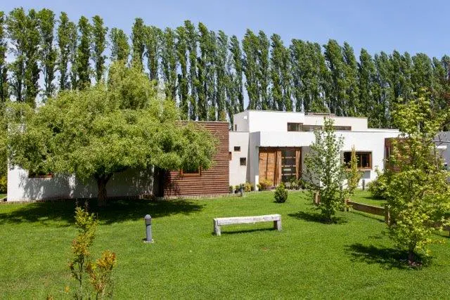 Modern white house with wood accents, surrounded by green lawn, trees, and a bench under a blue sky.