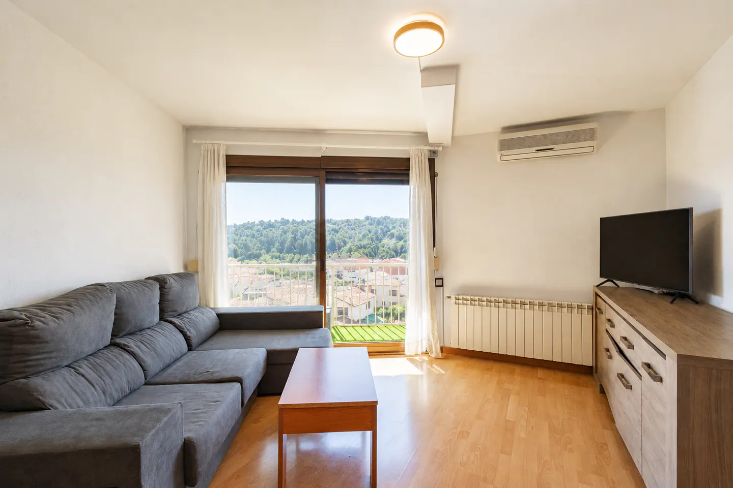 Bright living room with gray sectional sofa, wood floors, TV, and sliding glass doors to a balcony with a view.