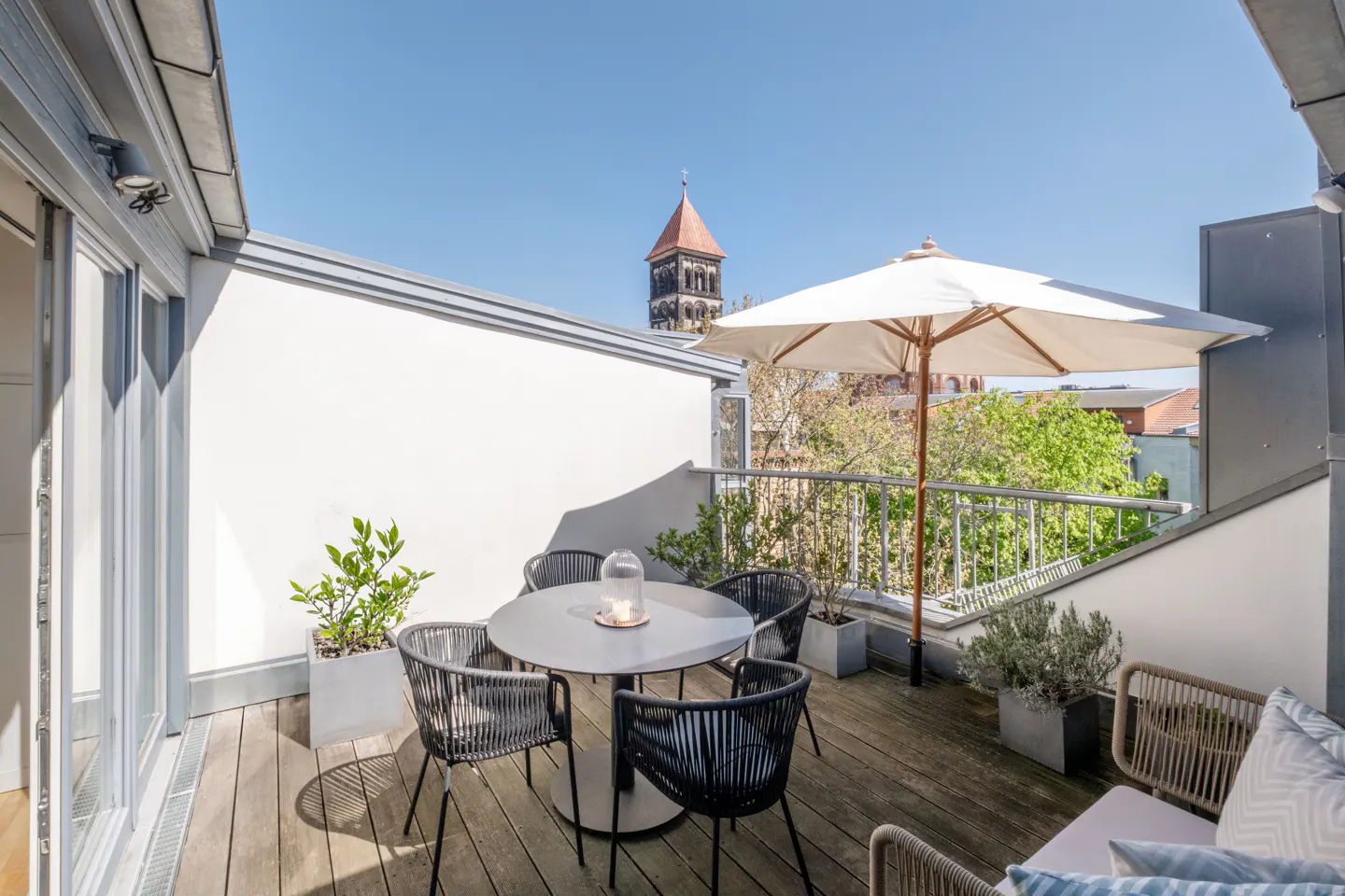 Outdoor patio with a round table, four chairs, and a white umbrella. A church tower is visible in the background.