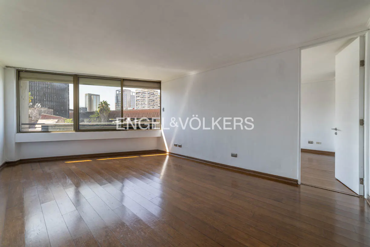 Empty room with hardwood floors, white walls, and large windows showing city views. A doorway leads to another room. Engel & Völkers logo is visible.