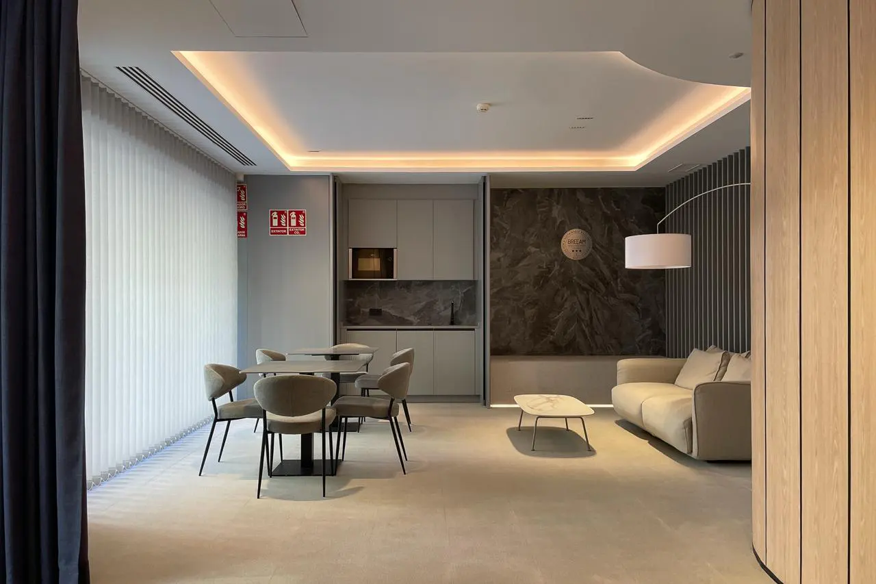 Modern office lounge with tables, chairs, a sofa, and a kitchenette. The room has a neutral color palette and recessed lighting.