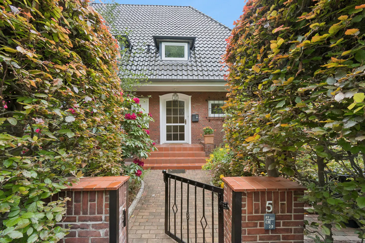 Brick house with a white front door, tiled steps, and a gated brick path framed by lush green and autumn-colored hedges.