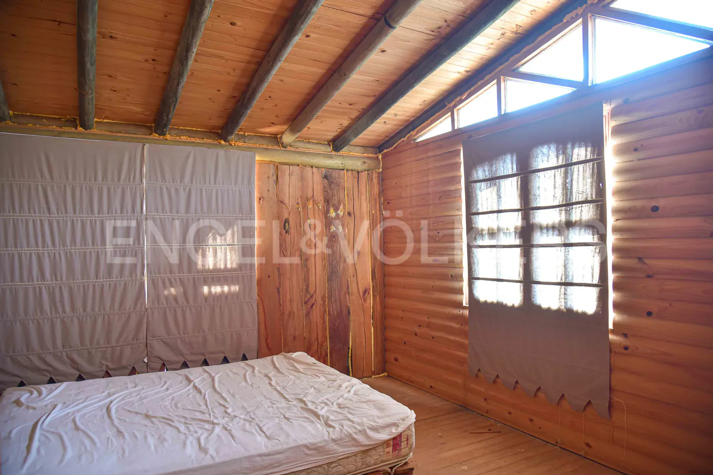 Bedroom with a white bed, wooden walls, and a window with a curtain. The ceiling is also wooden with dark beams.