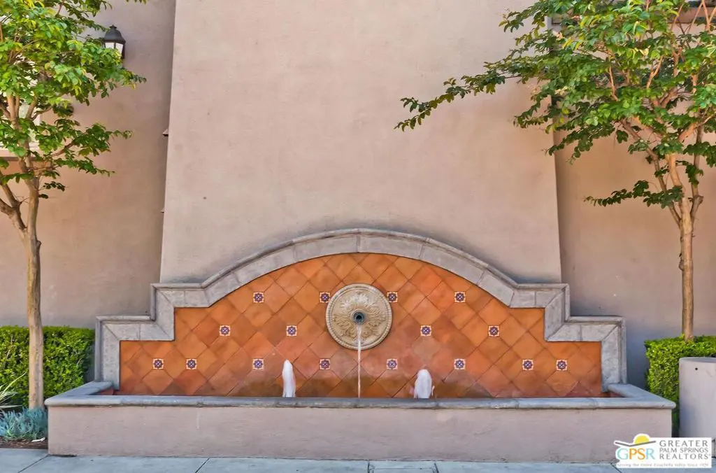 Outdoor fountain with orange tile backdrop and two trees on either side. Water flows from a central medallion.