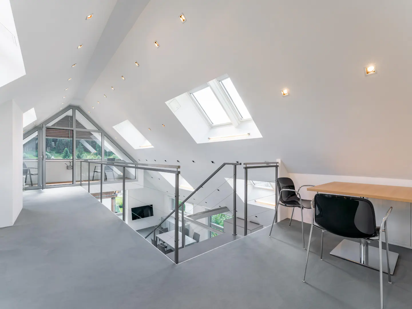 Bright, modern attic office space with skylights, glass railings, and a view to the floor below. Two black chairs sit by a light wood desk.