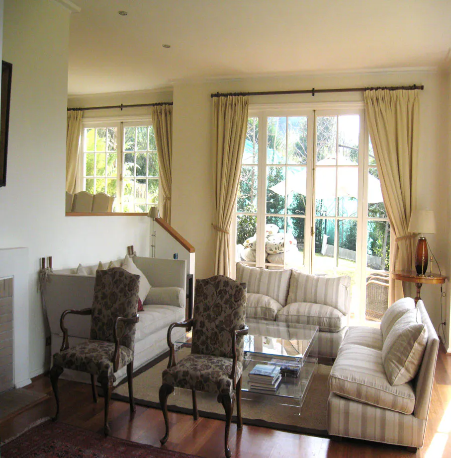 Living room with striped sofas, floral chairs, and large windows with cream curtains. A glass coffee table sits on a brown rug.