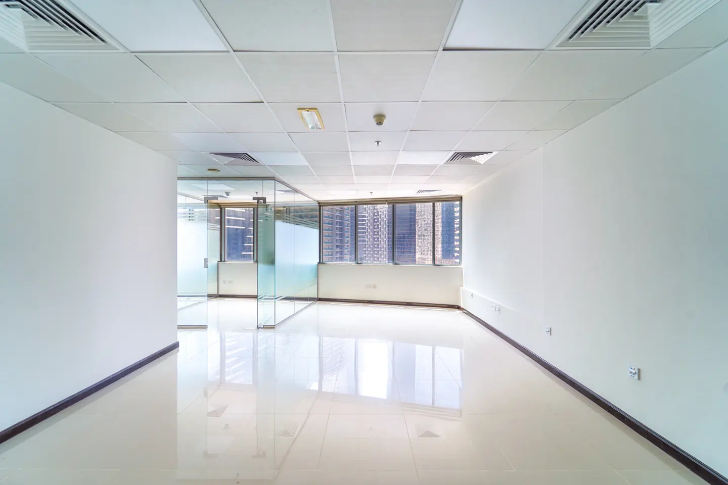 Empty office space with white walls, tile floor, and glass-walled rooms. Windows show a city skyline.