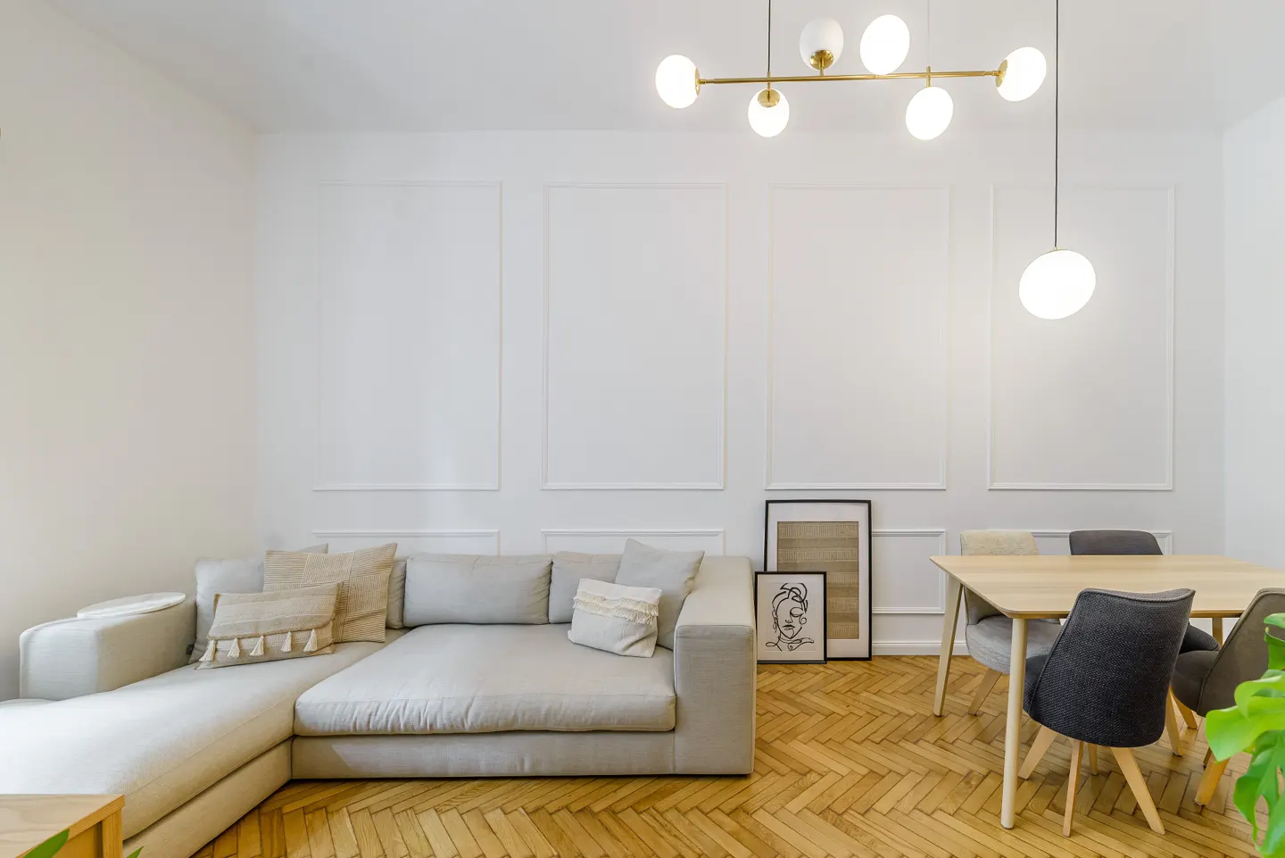 Modern living room with a beige sectional sofa, wooden dining table, herringbone floors, and white paneled walls.