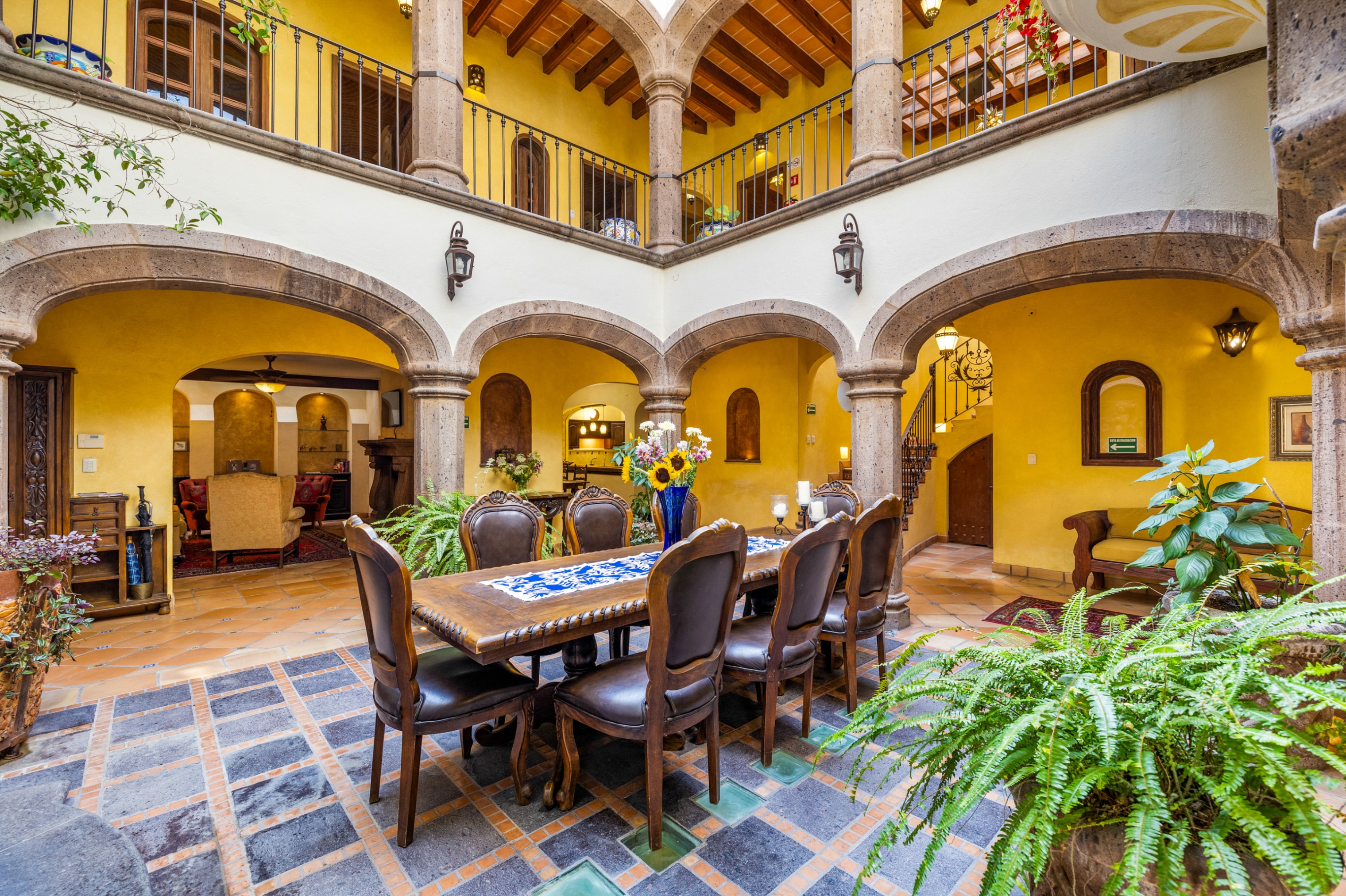 Interior courtyard with yellow walls, stone arches, and a long wooden table with brown leather chairs. Balcony above, plants around.