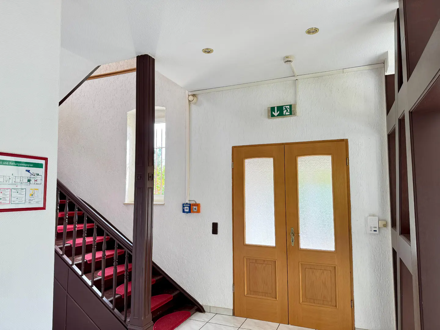 Interior view of a building with a staircase with red carpet, and a wooden door with a green exit sign above it.