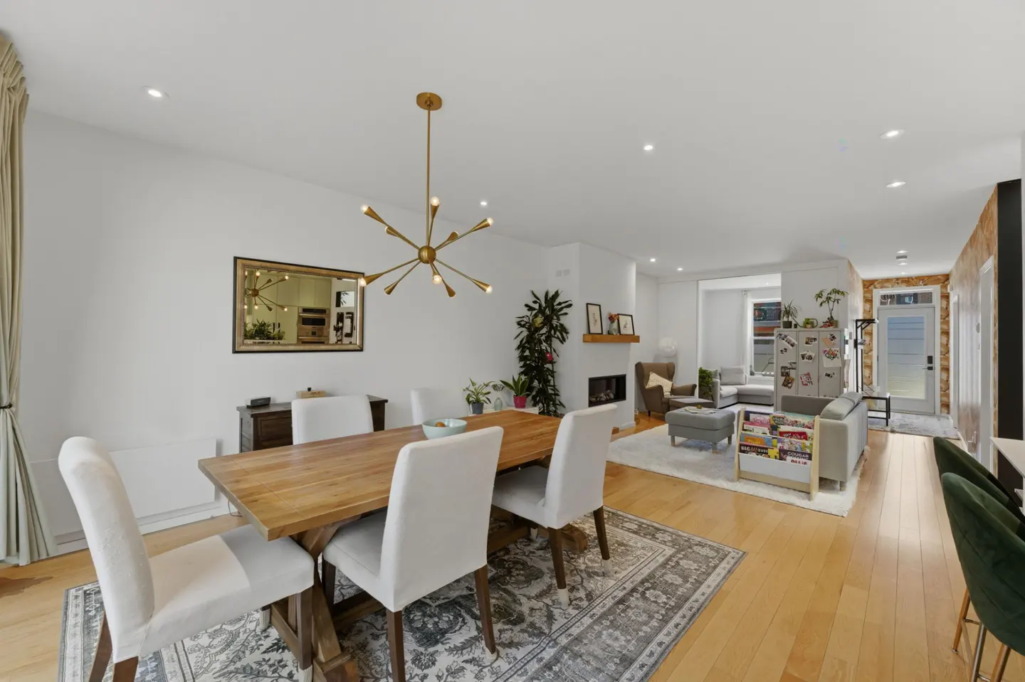 Bright, open-concept home with wood floors. Dining area with wood table, white chairs, and gold chandelier. Living room with fireplace and gray sofa.
