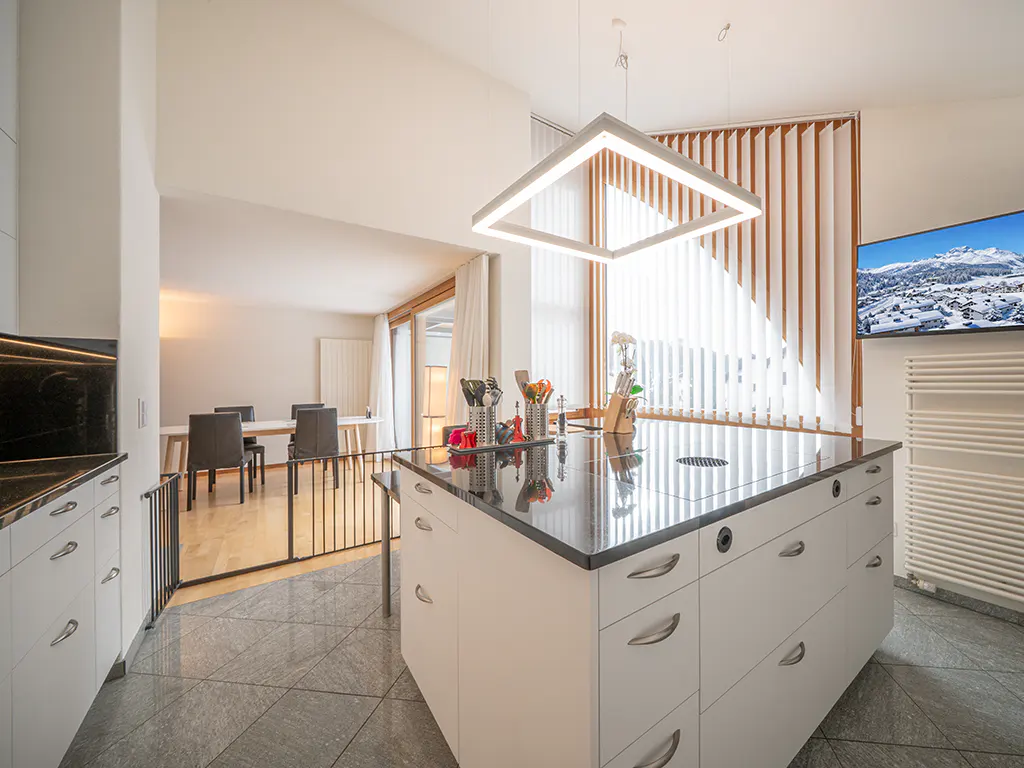 A modern kitchen with a white island, black countertop, and square light fixture. A dining area is visible in the background.