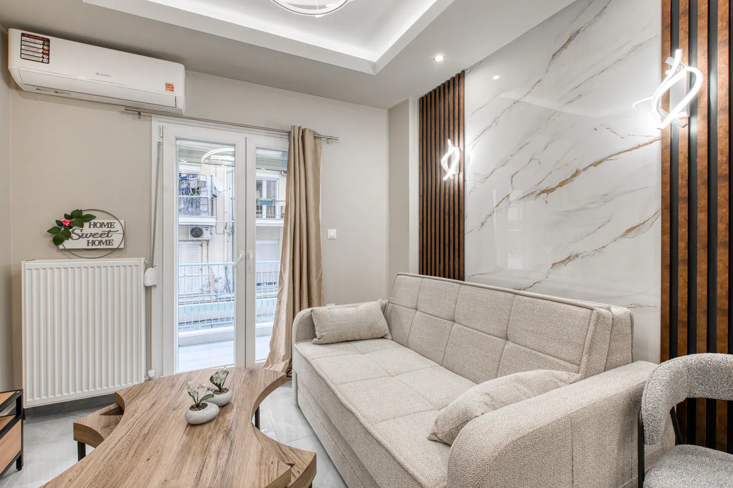 Living room with a beige sofa, wood coffee table, and marble accent wall with modern sconces. Balcony doors let in natural light.