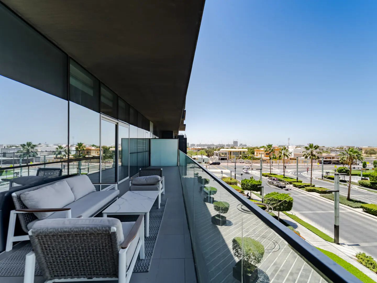 Balcony view with gray outdoor furniture, a white table, and a glass railing overlooking a street with palm trees on a sunny day.