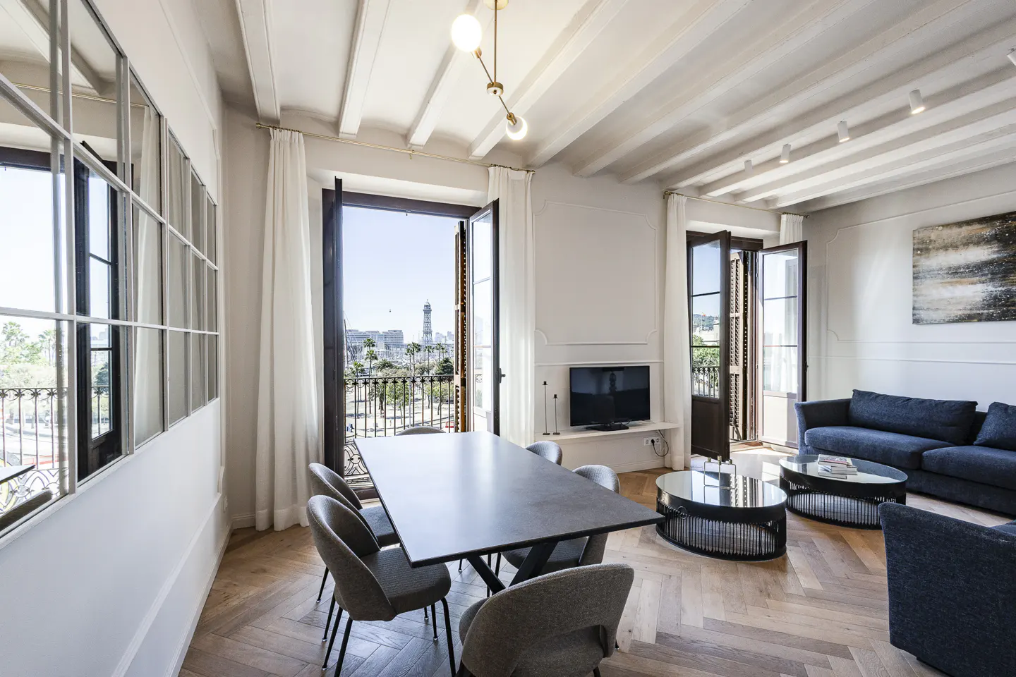 Bright living room with herringbone floors, a dining table, and a blue sofa. Balcony doors open to a city view.