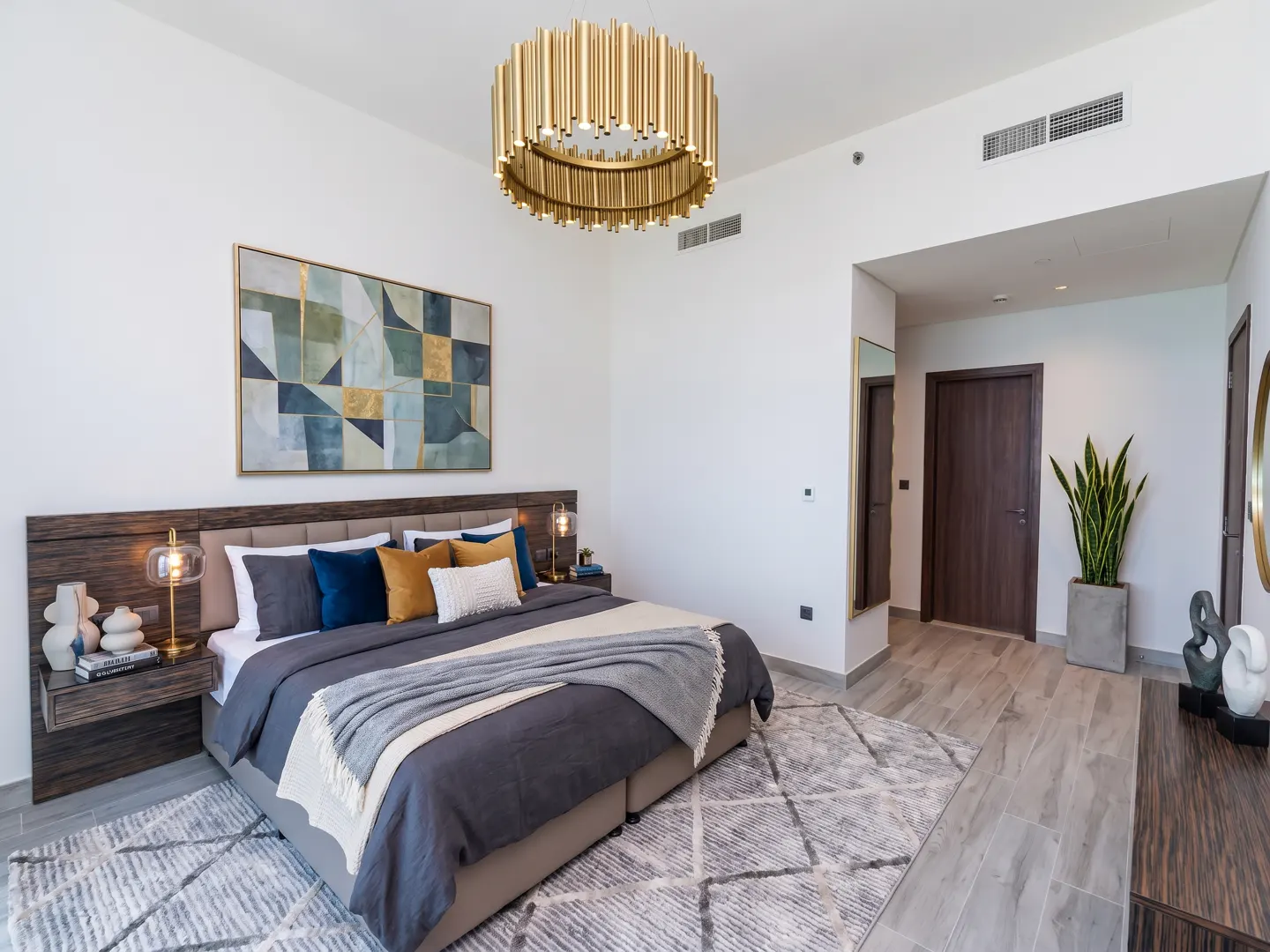 A modern bedroom with a king-size bed, gray duvet, and colorful pillows. A gold chandelier hangs above, and abstract art adorns the white walls.