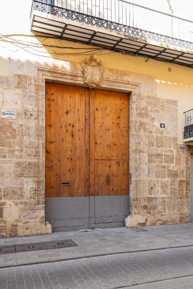 A large, weathered wooden door set in a stone facade with a gray base, under a balcony with black ironwork.