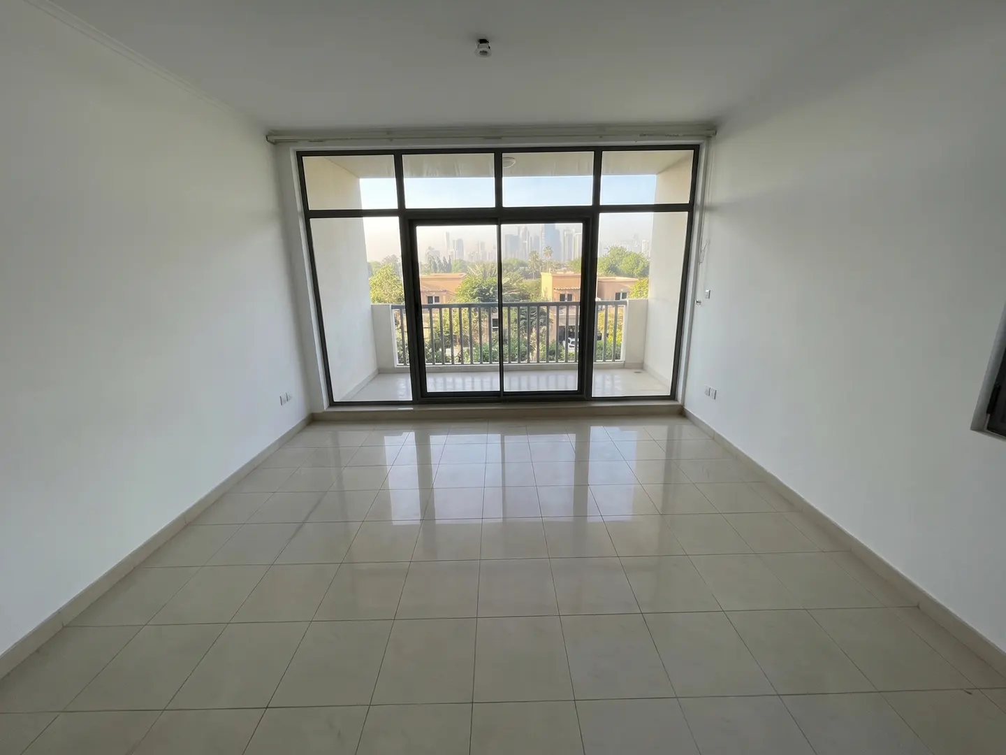 Empty room with white walls and beige tiled floor. Sliding glass doors lead to a balcony with a view of buildings and trees.
