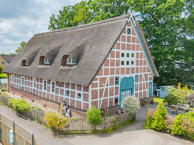 Traditional half-timbered house with a thatched roof, brick walls, and teal accents, surrounded by a garden and trees.