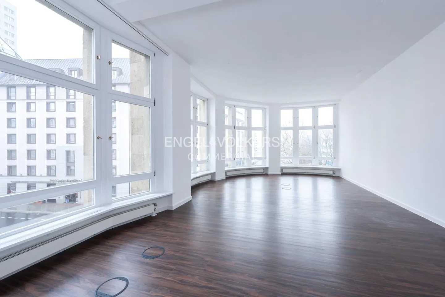 Bright, empty room with dark wood floors and white walls. Large windows offer a city view. "Engel & Volkers Commercial" logo visible.