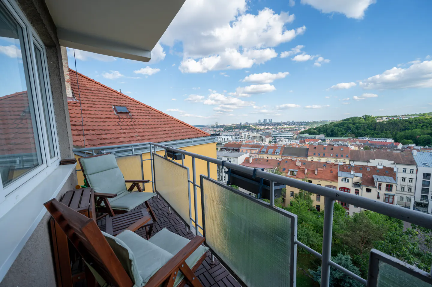 Balcony view with two chairs, a small table, and a cityscape in the background under a blue sky with white clouds.