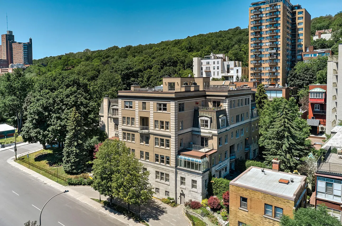 A tan brick apartment building with many windows, surrounded by green trees and other buildings.