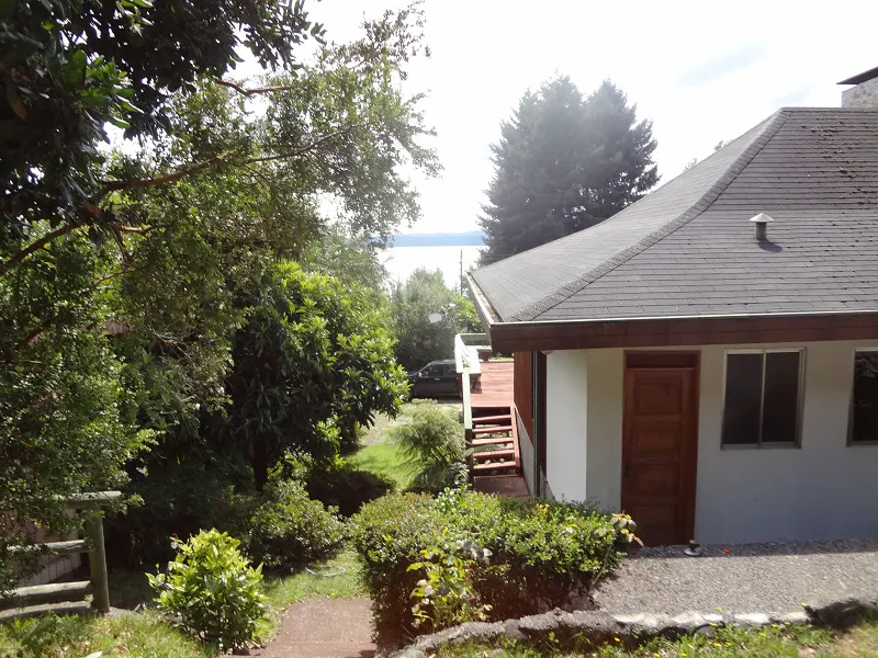 Exterior view of a white house with a gray roof, brown trim, and a wooden door, surrounded by lush greenery and a glimpse of a lake in the distance.
