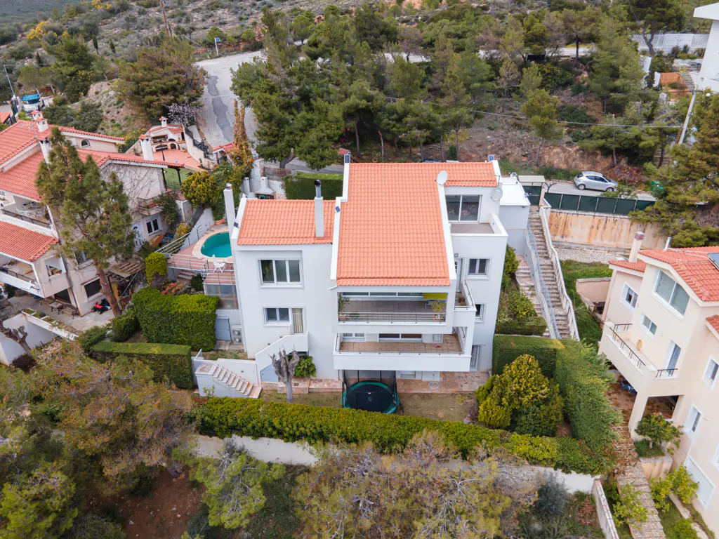 Aerial view of a white, multi-story house with a red tile roof, surrounded by green trees and bushes on a hillside.
