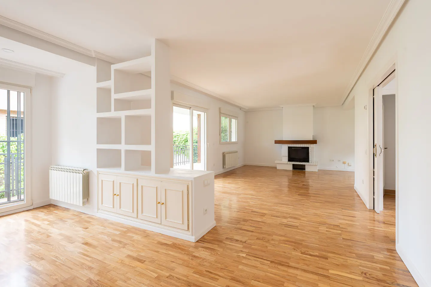 Bright, empty living room with hardwood floors, white walls, and a fireplace. A white bookcase divides the space. Balcony access is visible.