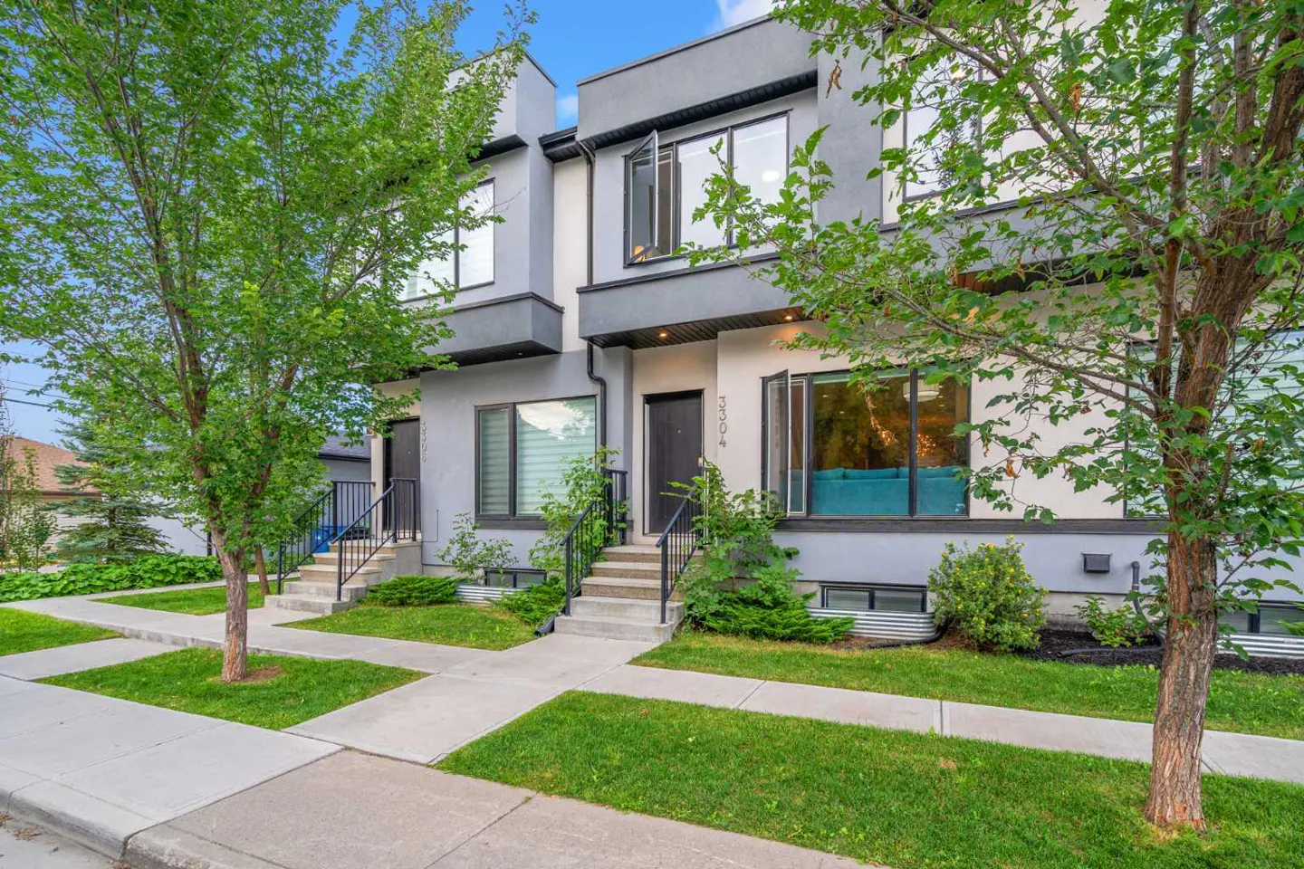 Exterior view of two-story gray townhouses with black doors, green lawns, and trees under a blue sky.
