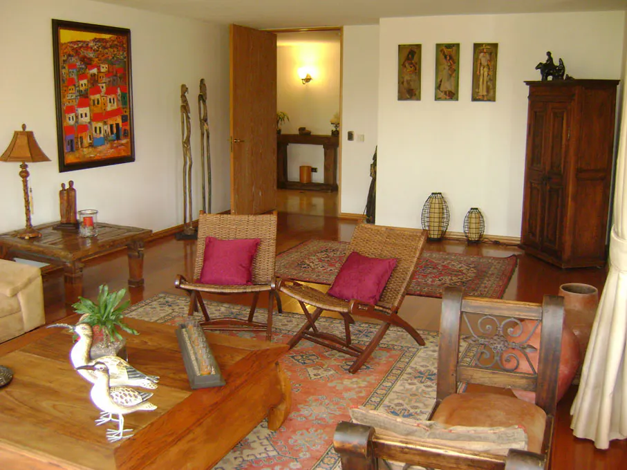 Living room with hardwood floors, wicker chairs with red pillows, and a large painting of a village scene.