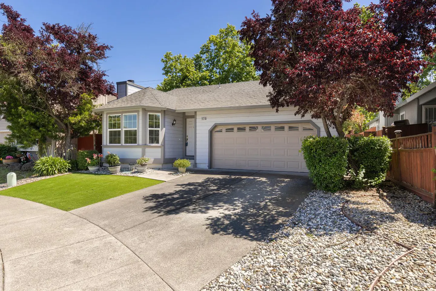 A single-story, light gray house with a beige garage door, green lawn, and trees under a blue sky.