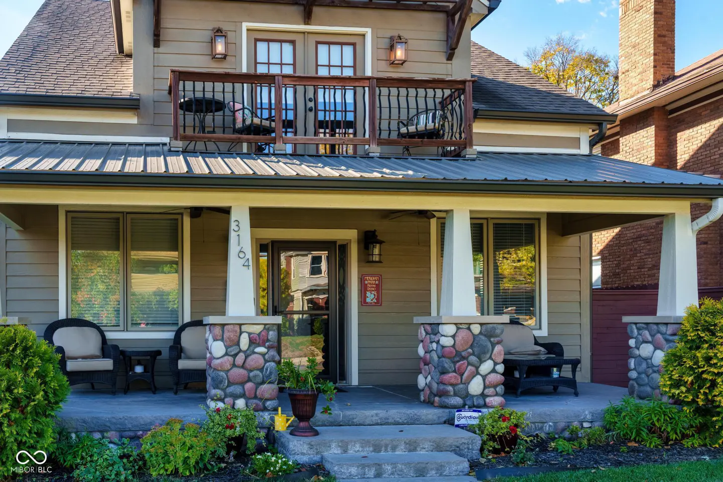 Two-story house with a porch and balcony. The porch has stone pillars and wicker furniture. The balcony has a metal railing and chairs. The house number is 3164.