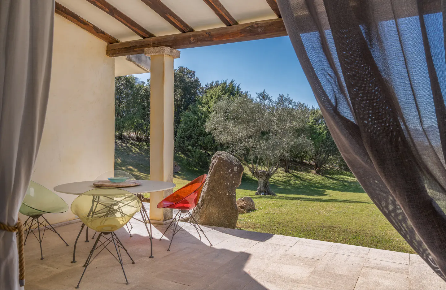 Outdoor patio with round table, colorful chairs, and view of green lawn with trees. Gray curtains frame the right side of the image.