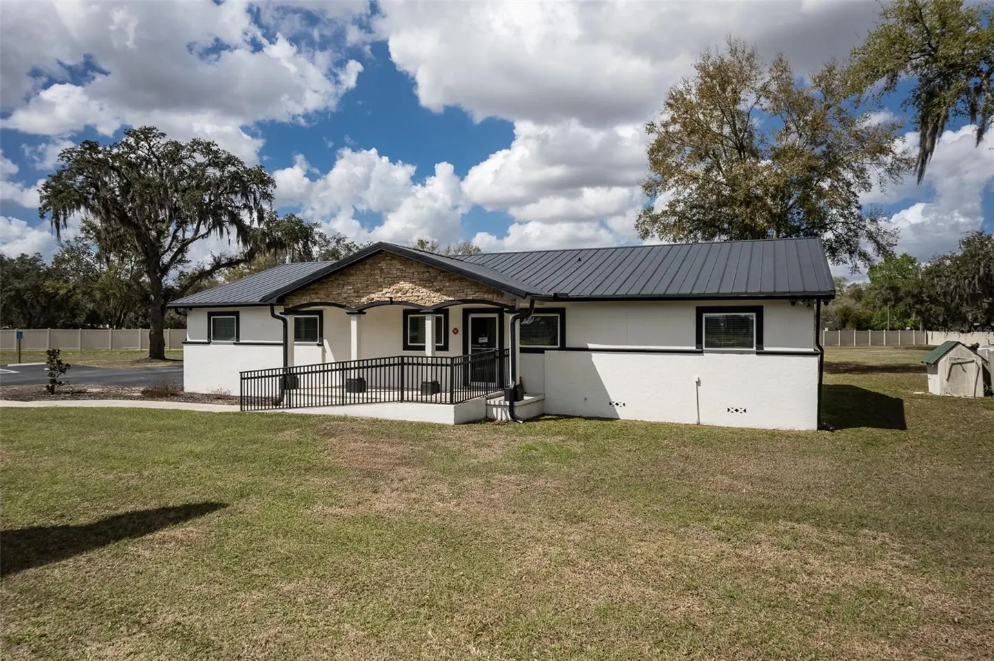 A white, one-story building with a gray metal roof and stone accents, surrounded by a green lawn and trees.