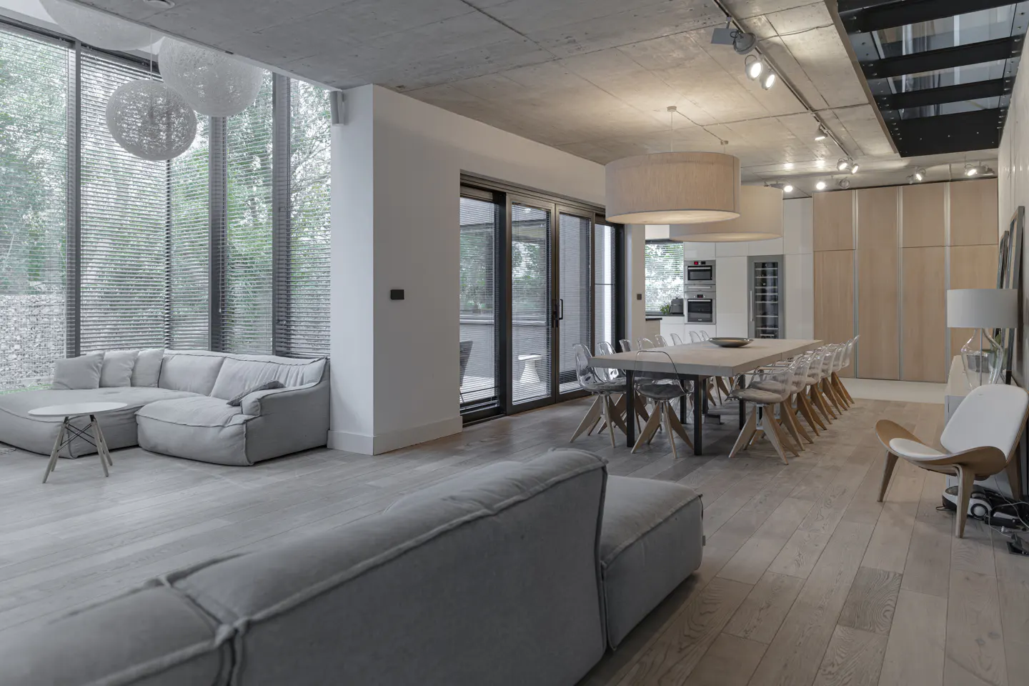 Open-concept living space with gray sofas, a long dining table with clear chairs, and light wood floors. Large windows offer natural light.