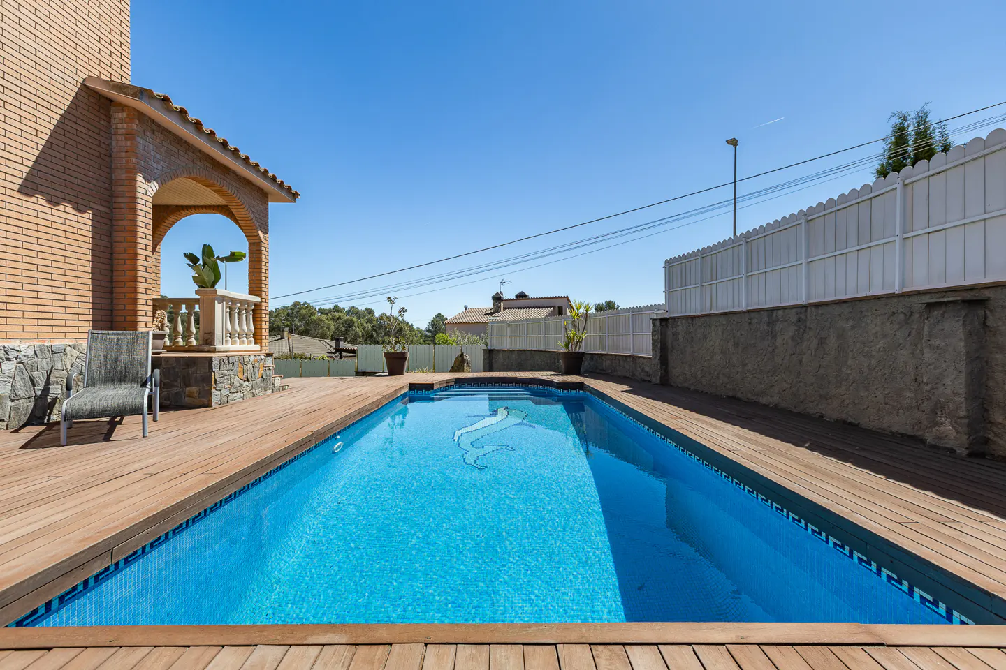 A backyard pool with blue tile and a dolphin mosaic is surrounded by a wooden deck. A brick house and white fence are in the background.