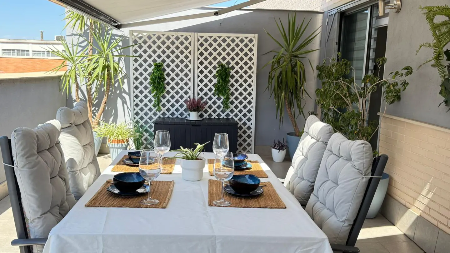 Outdoor dining area with a white tablecloth, place settings, and cushioned chairs. Plants and a white lattice screen decorate the patio.