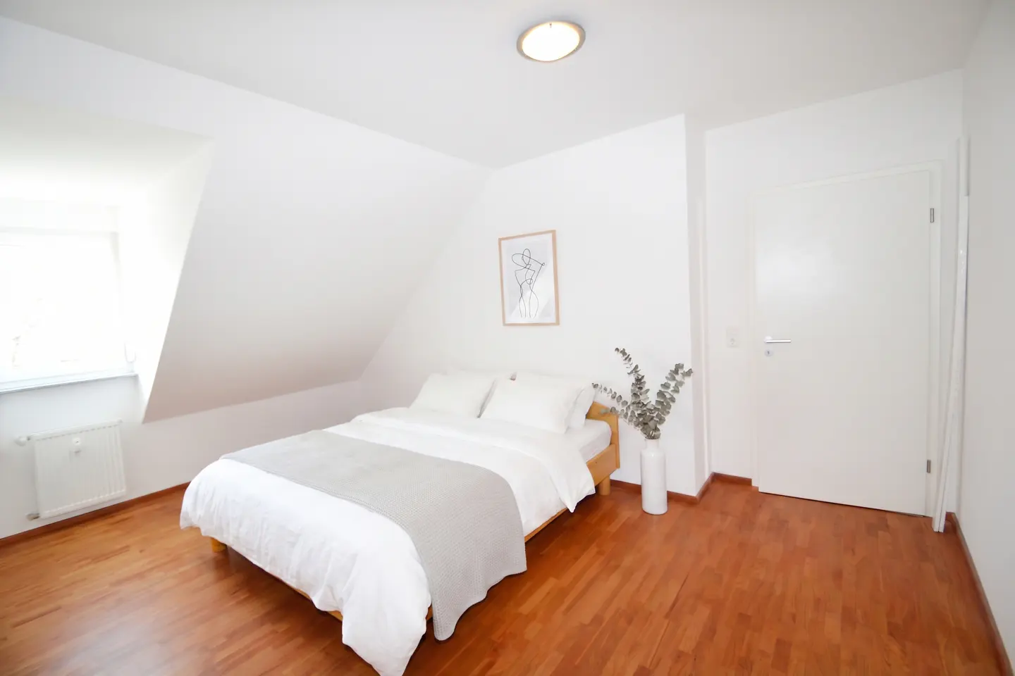 Bright bedroom with white walls, wood floor, and a bed with white linens and a gray throw. A framed art print hangs above the bed.