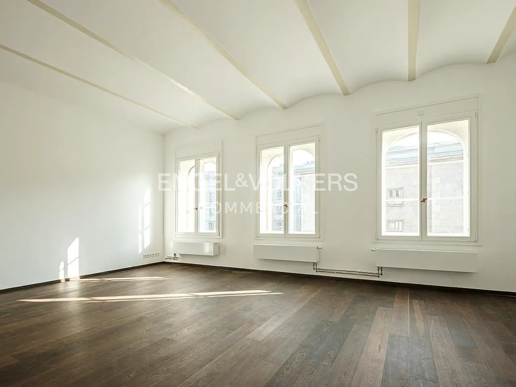 Bright, empty room with dark wood floors, white walls, and three arched windows. Beamed ceiling and baseboard heaters.