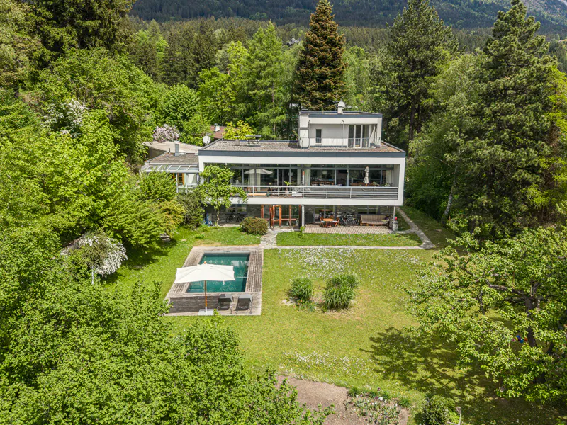 Aerial view of a modern white house with a pool, surrounded by lush green trees.