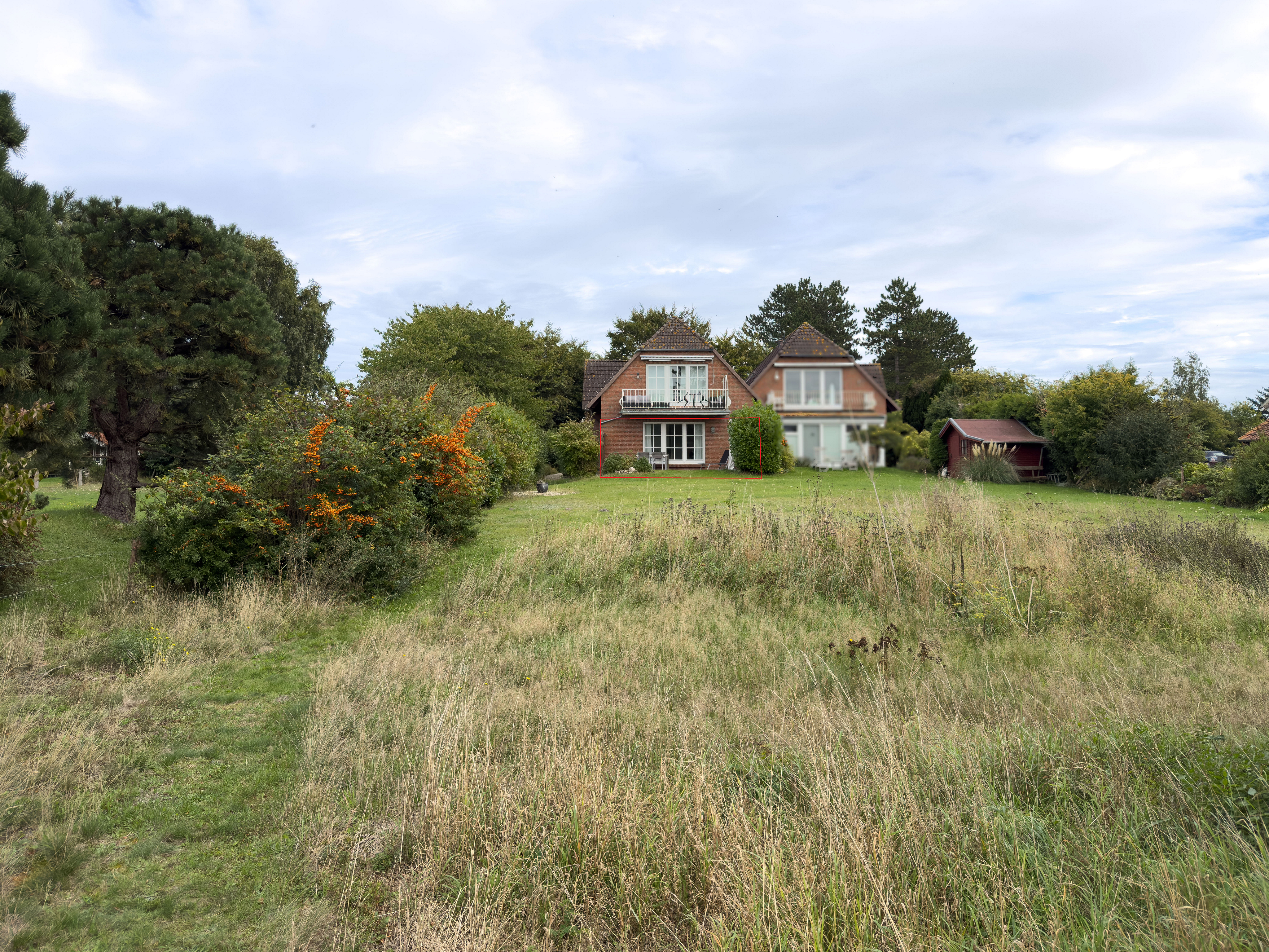 A two-story brick house with balconies sits behind a large, grassy field surrounded by trees and shrubs under a cloudy sky. [AI-generated text]