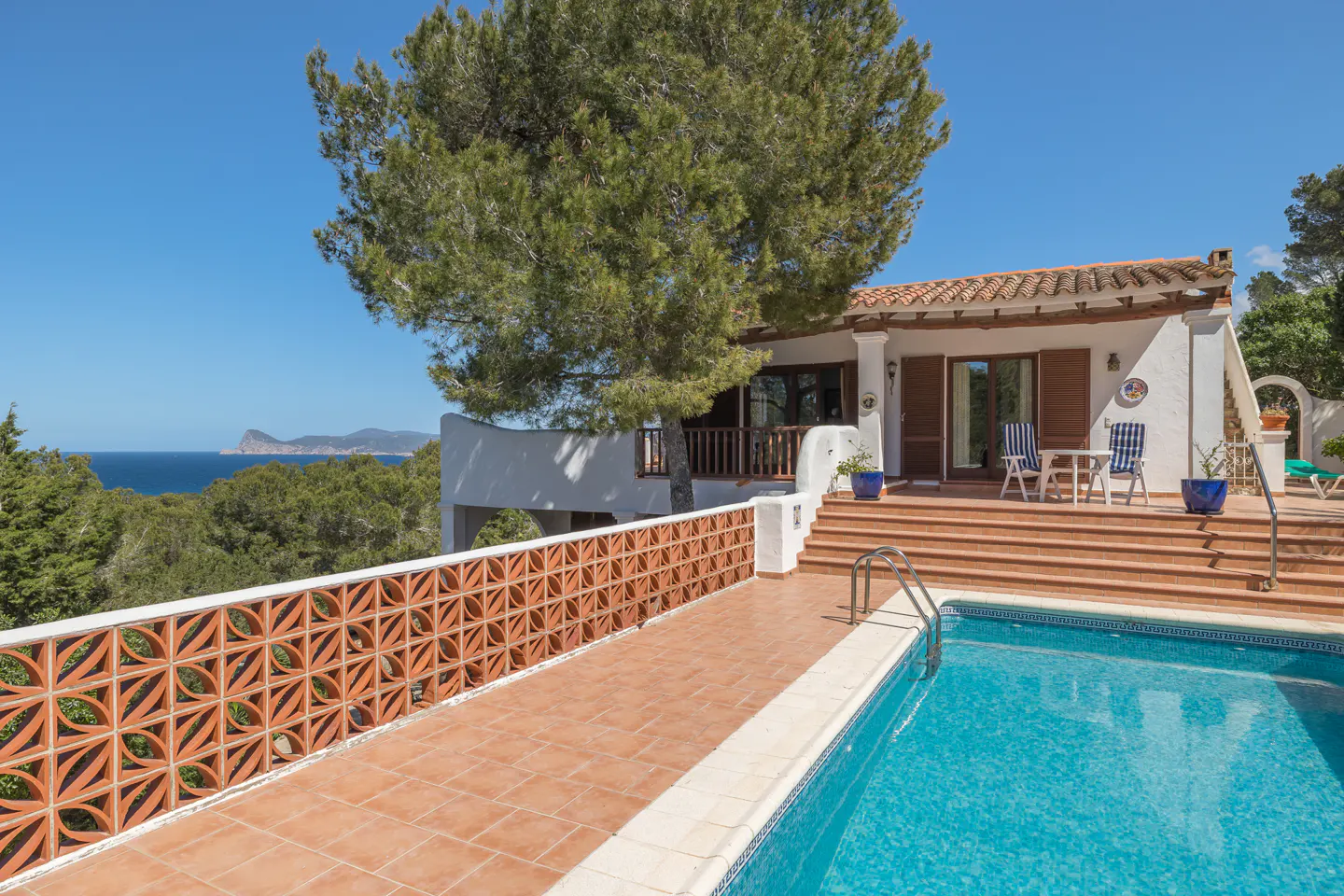 Exterior view of a white villa with a pool, terracotta patio, and ocean view under a clear blue sky.
