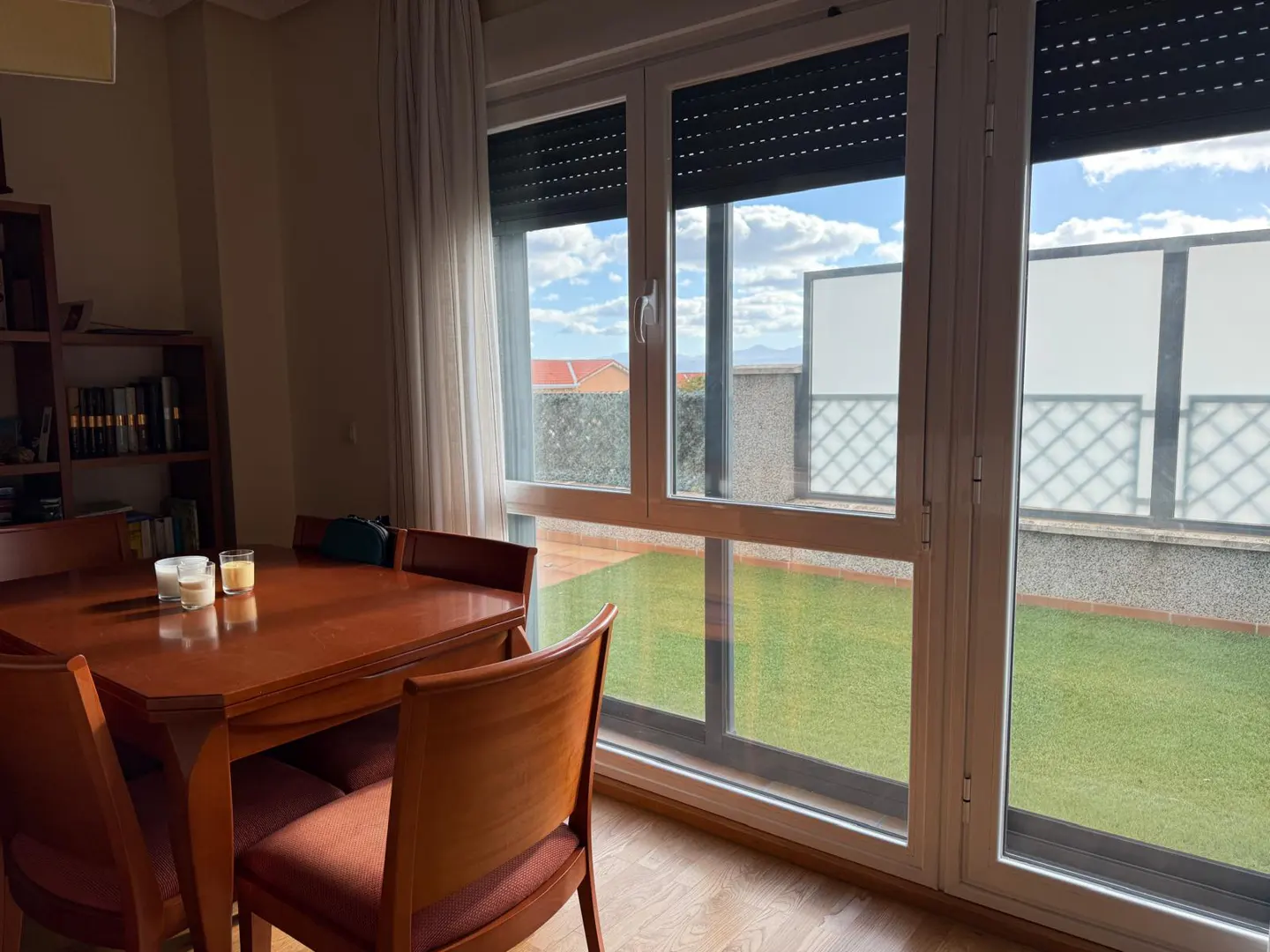 Dining room with a wooden table, chairs, and a large window overlooking a green terrace and blue sky.