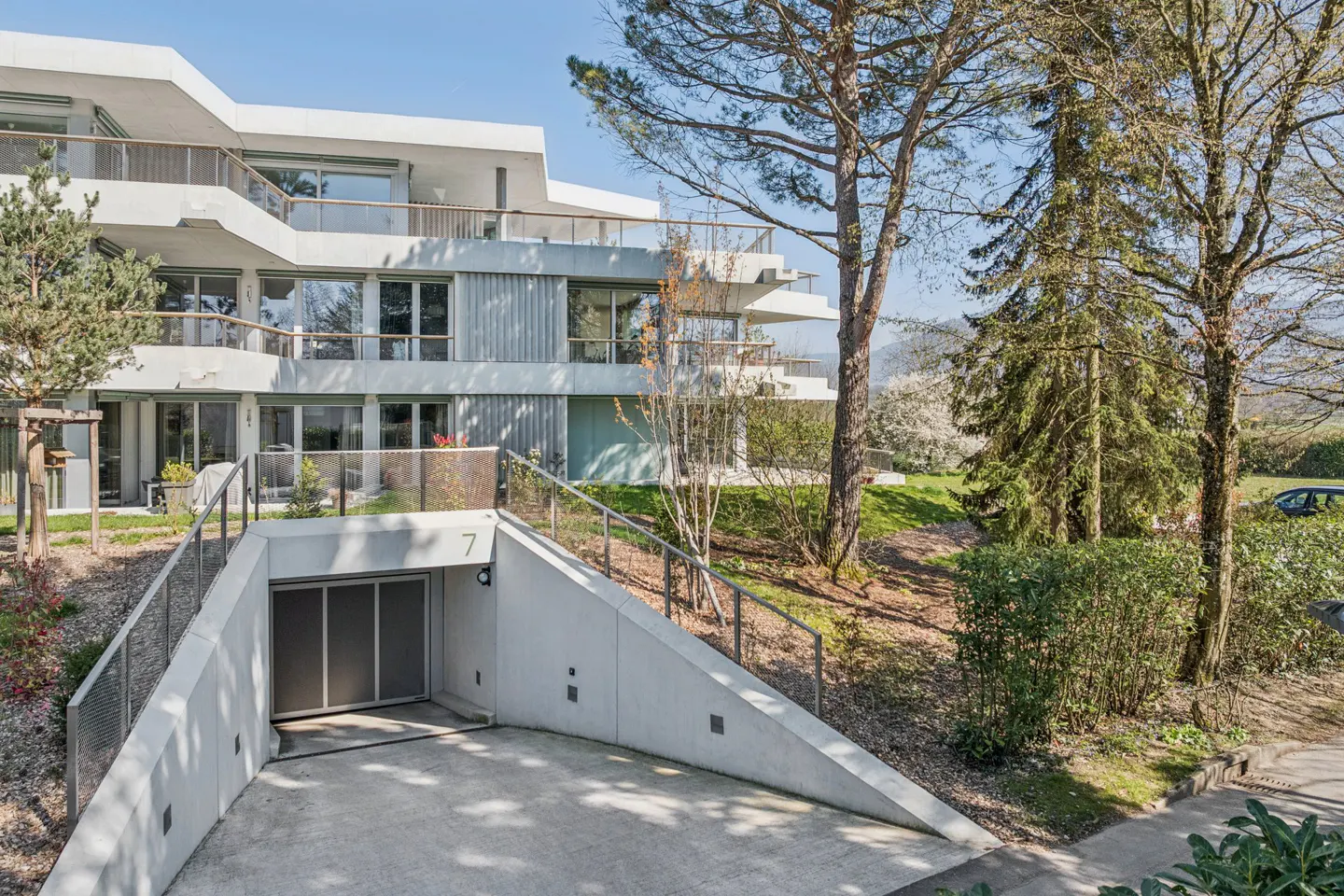 Modern white apartment building with balconies and a concrete garage entrance marked with the number 7. Trees and greenery surround the building.