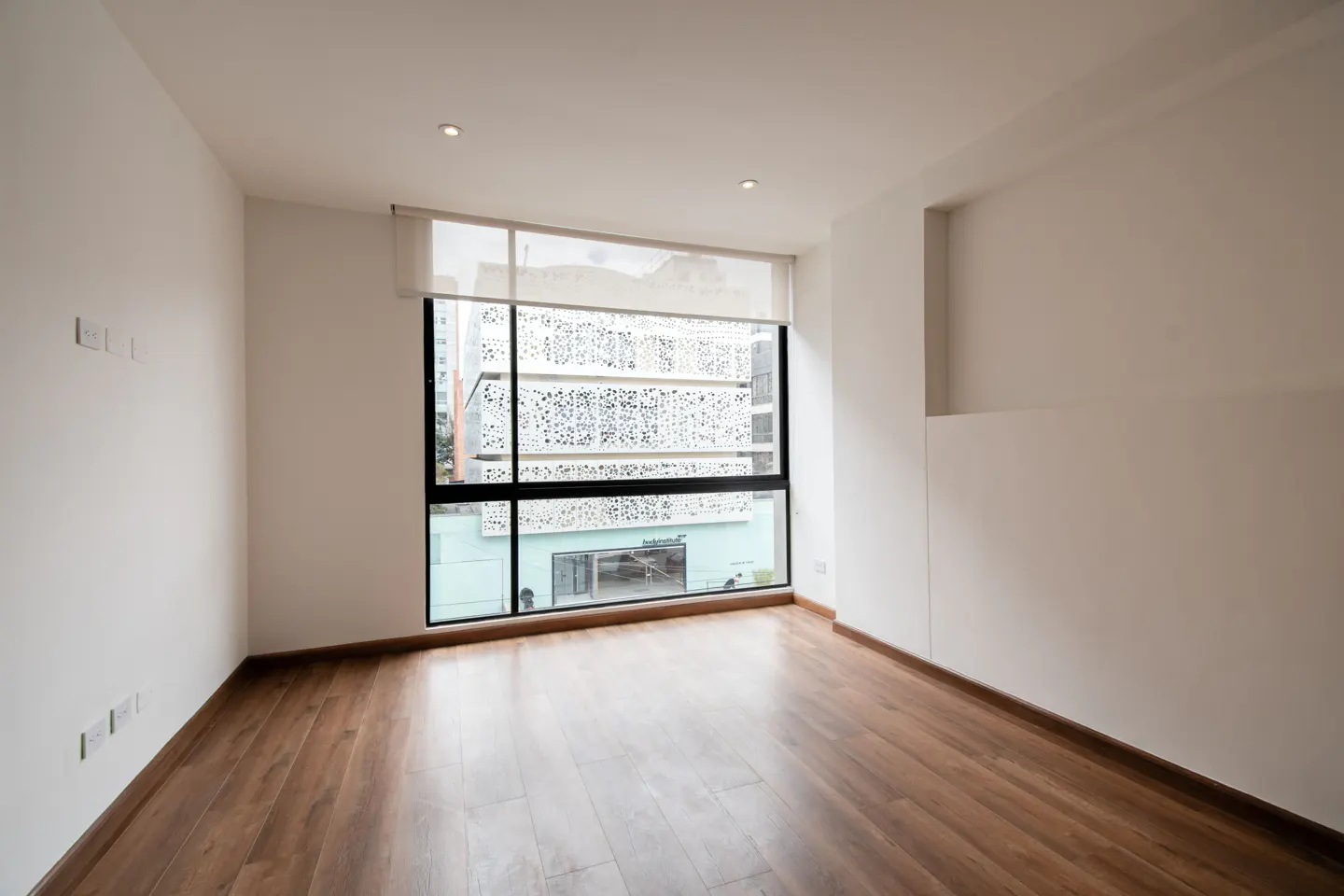 Empty room with wood floors, white walls, and a large window. Outside, a building with a patterned facade is visible.