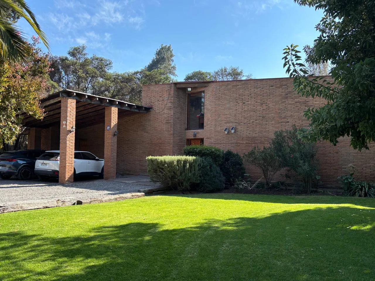 Brick house with a carport and green lawn. Two cars are parked under the carport. Bushes are in front of the house.