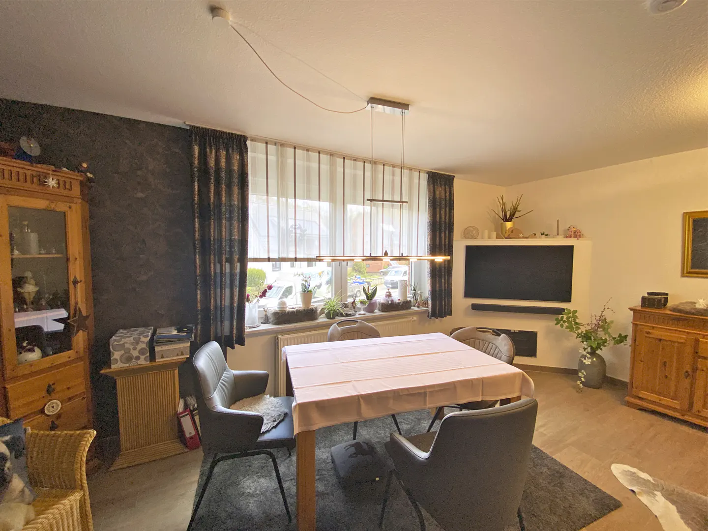 A dining room with a table, chairs, and a window with curtains. A TV and wooden cabinet are in the background.