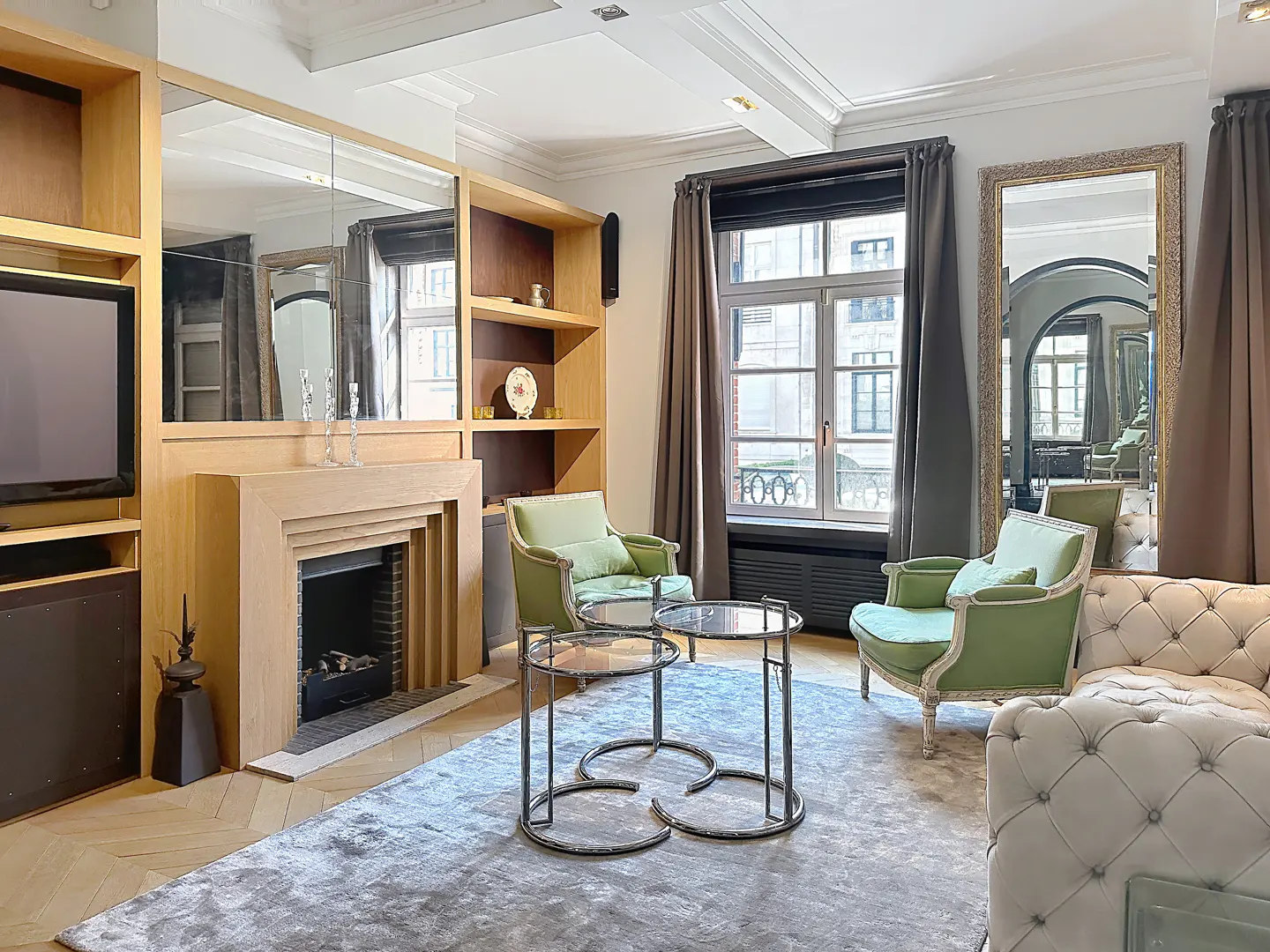 Living room with fireplace, TV, and shelves. Two green armchairs face a window with gray curtains. A gray rug covers the herringbone wood floor.