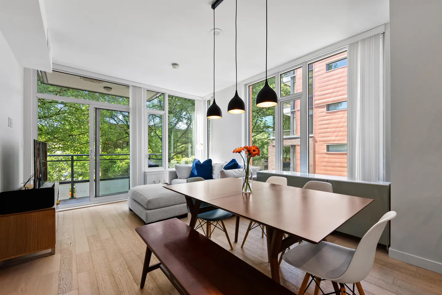 Modern dining area with a wooden table, bench, and chairs, featuring large windows, pendant lights, and a cozy sofa.